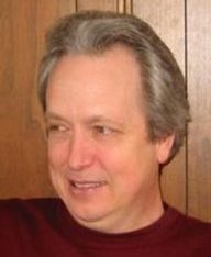 David Griffeath, smiling, with grey shoulder-length hair and a dark red jumper, in front of a wooden door
